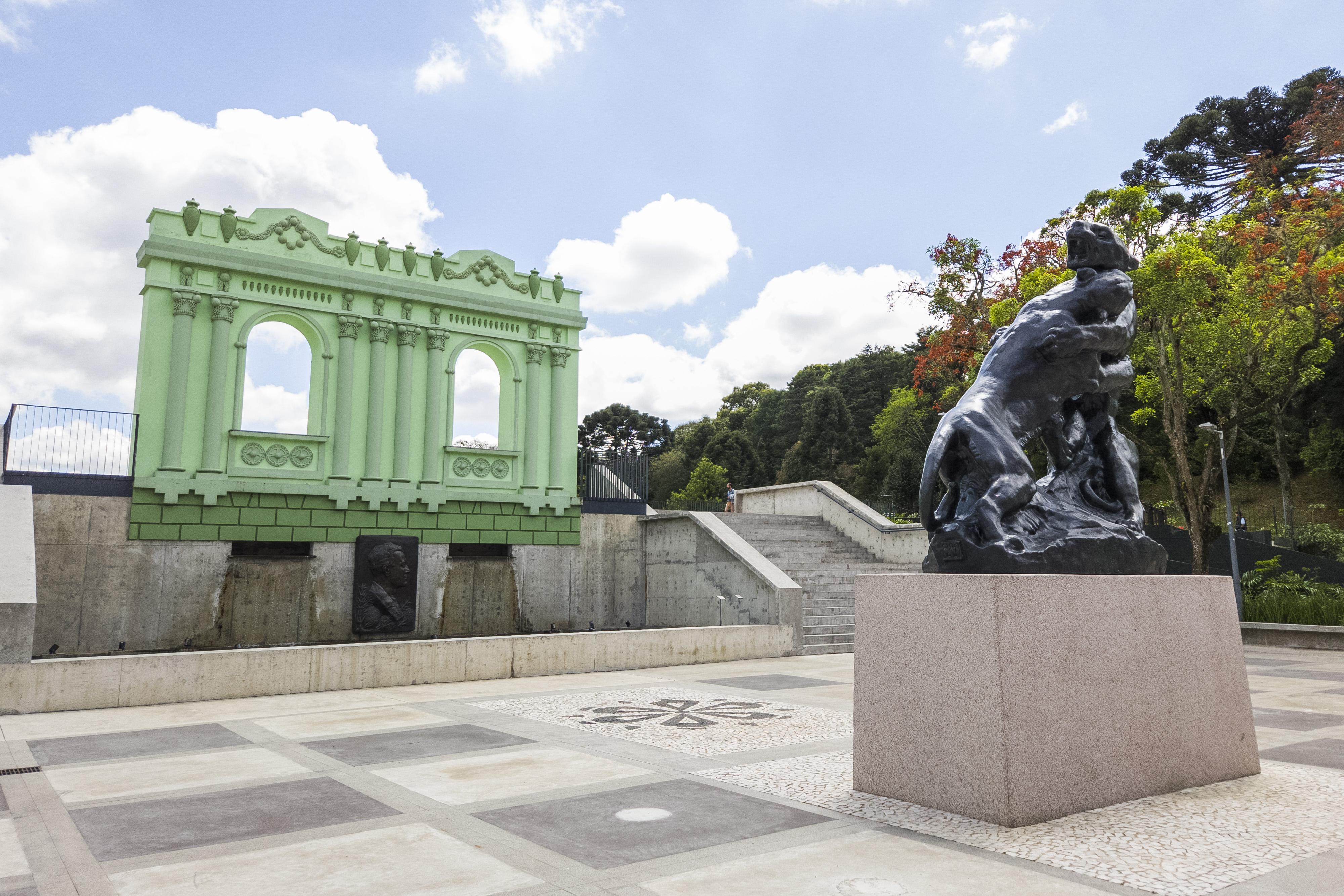 Vista externa do Memorial Paranista, no Parque São Lourenço, em Curitiba, com uma grande escultura de onças em bronze, estrutura arquitetônica verde, escadarias e área de visitação ao ar livre.