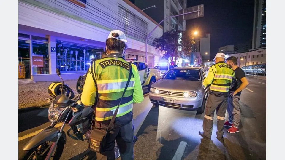 Imagem ilustrativa de agentes realizando fiscalização noturna de trânsito com veículos e cones de sinalização.
