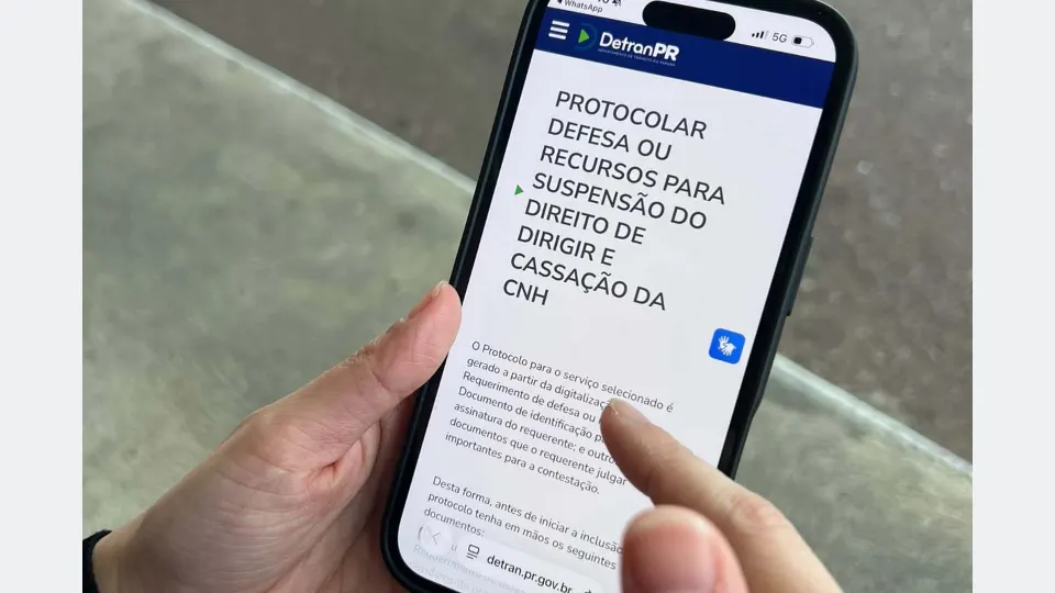 Pessoa segurando celular com página do Detran-PR destinada à apresentação de defesa e recursos relacionados à suspensão ou cassação da CNH.