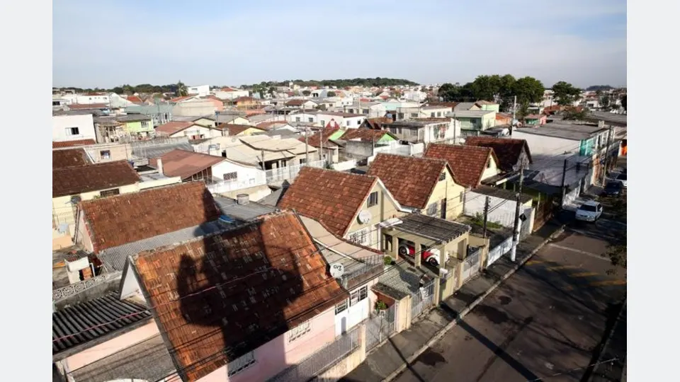 Vista panorâmica da Vila Nossa Senhora da Luz, na CIC, em Curitiba, com casas de telhados inclinados, ruas residenciais e diversas moradias ao fundo, mostrando a transformação do conjunto habitacional ao longo de seis décadas.