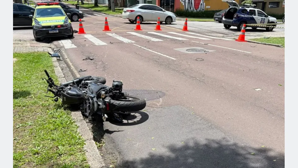 Motocicleta preta caída próximo à calçada após acidente no bairro Rebouças, em Curitiba. Ao fundo, viaturas de trânsito e da Guarda Municipal, além de cones de sinalização isolando o cruzamento.