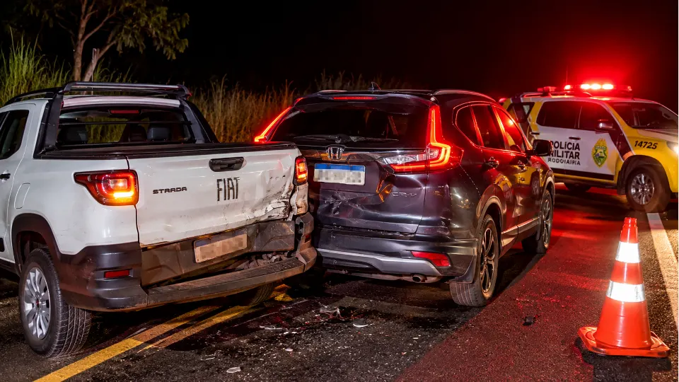 Ilustração de colisão entre uma Fiat Strada e um Honda CR-V, com viatura policial e sinalização noturna em rodovia.