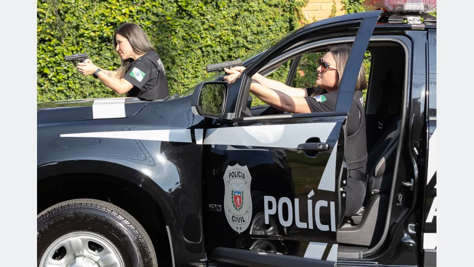 Duas policiais civis do Paraná participam de treinamento com armas de fogo ao lado de uma viatura preta da corporação, em ambiente externo.