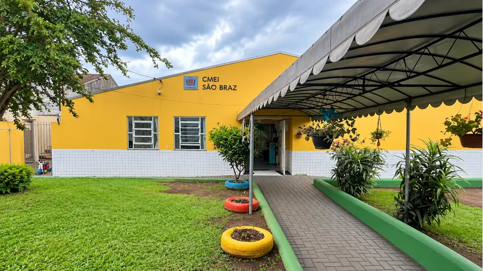 Fachada amarela do CMEI São Braz, em Curitiba, com entrada coberta, jardim e acesso principal da unidade de educação infantil.