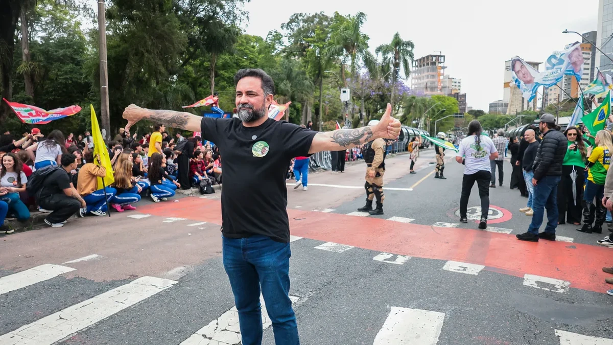 Homem faz gestos com os braços em frente a manifestantes reunidos em uma avenida de Curitiba, com estudantes sentados e policiais ao fundo.