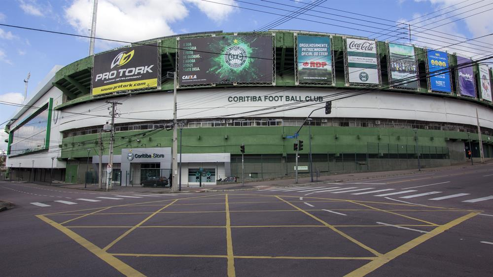 Vista externa do Estádio Couto Pereira, em Curitiba, com fachada verde e branca, identificação do Coritiba Foot Ball Club e cruzamento de ruas em frente ao estádio.