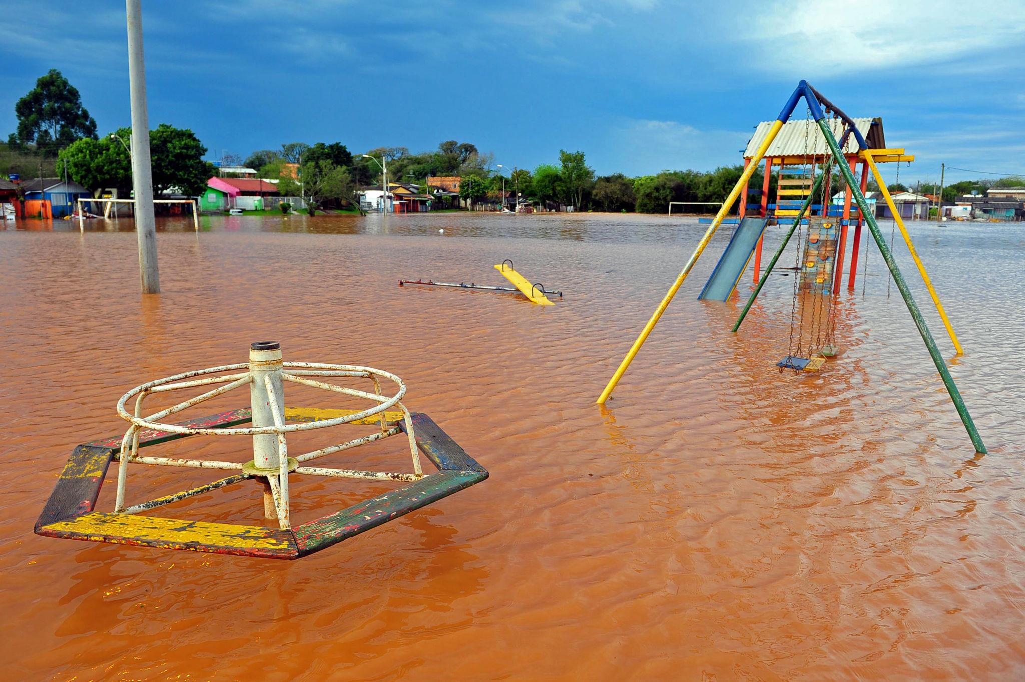 Parque infantil inundado por água barrenta, com balanços, escorregador e gira-gira parcialmente submersos.