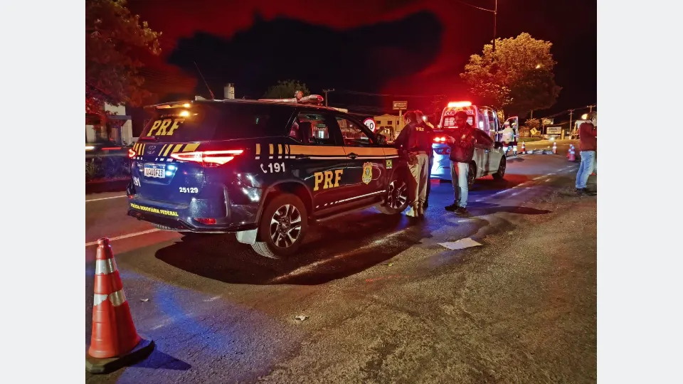 Viatura da Polícia Rodoviária Federal e ambulância em trecho de rodovia sinalizado com cones durante atendimento noturno.