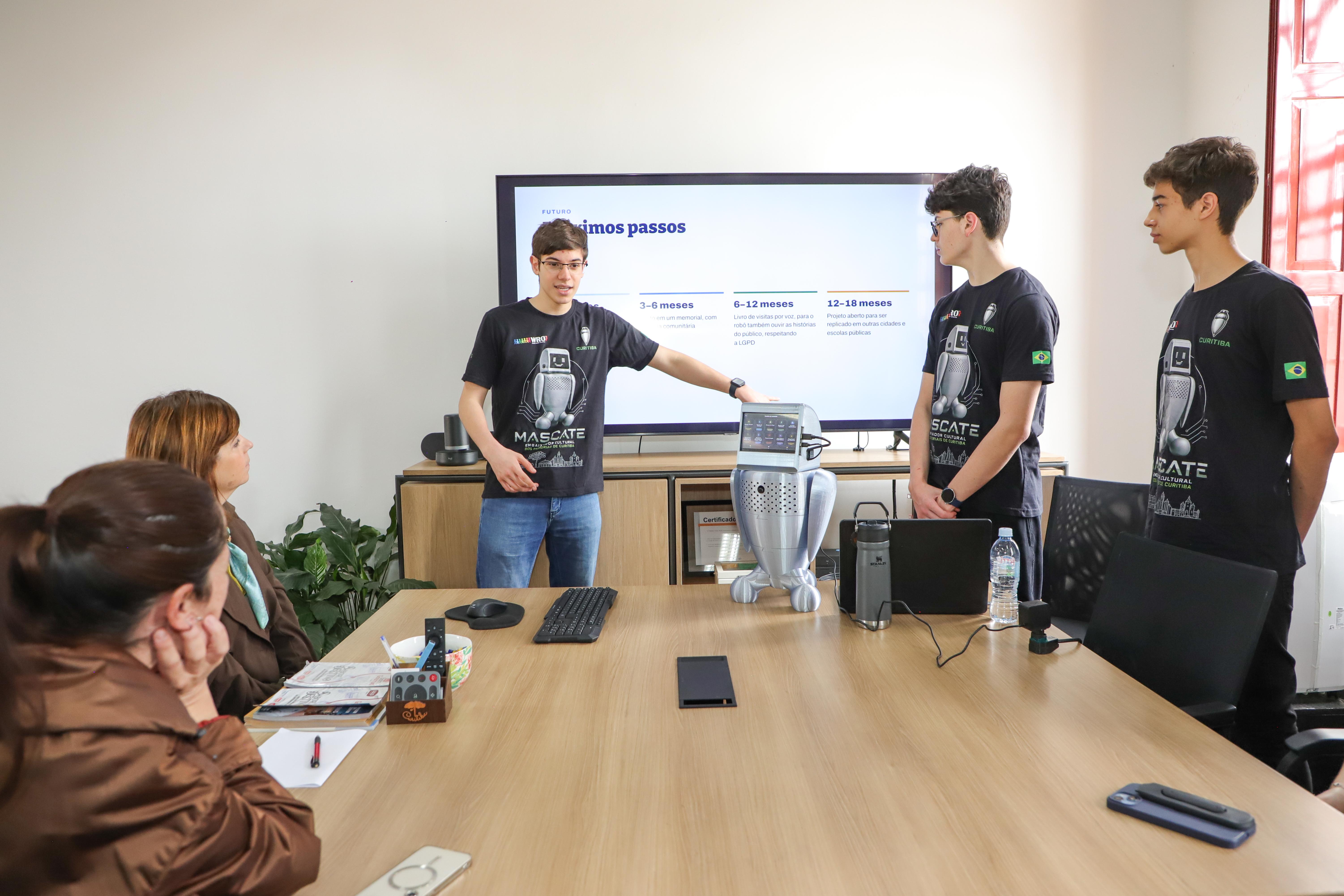 Estudante apresenta o funcionamento do robô Mascate diante de uma tela, acompanhado de outros integrantes da equipe.