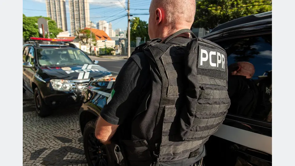 Policial civil do Paraná, usando colete tático com identificação PCPR, ao lado de viaturas policiais em uma rua urbana, durante atividade operacional.
