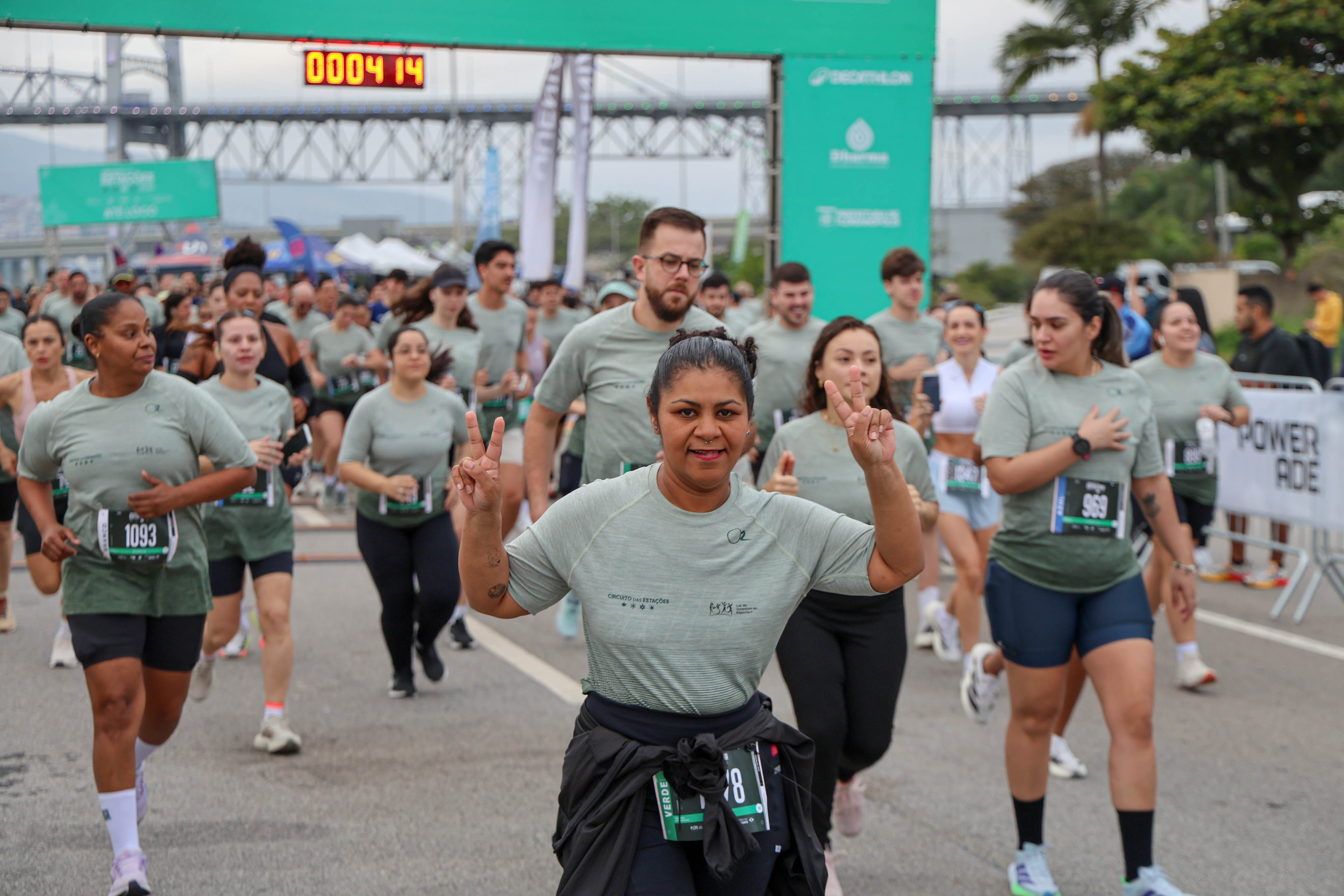 Corredores participam do Circuito das Estações, com atleta em primeiro plano fazendo sinal de vitória e outros participantes ao fundo.
