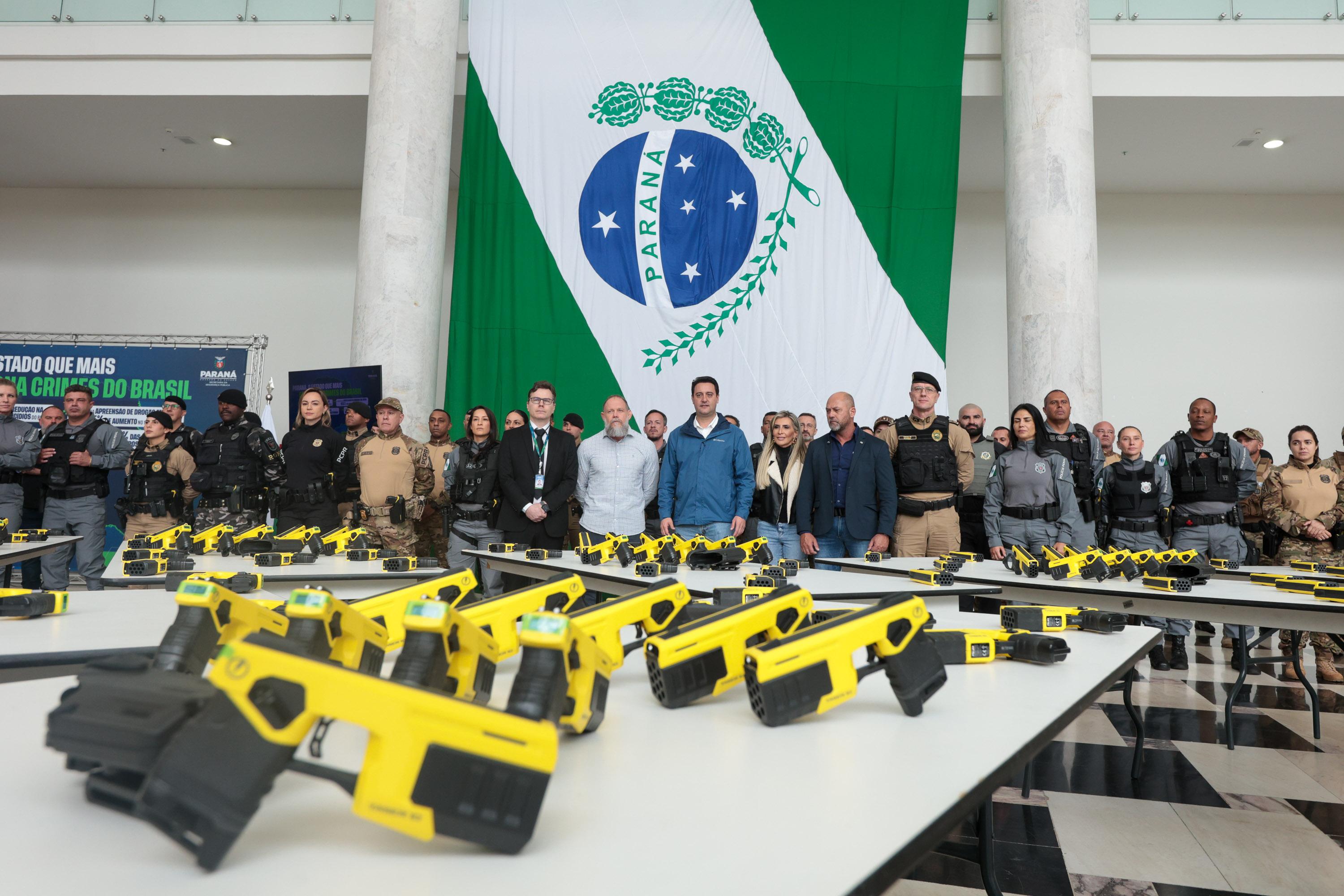 Dispositivos de incapacitação neuromuscular amarelos, espargidores de gás de pimenta e maletas de equipamentos dispostos sobre uma mesa, diante de painel com a identificação da Guarda Municipal de São José dos Pinhais.
