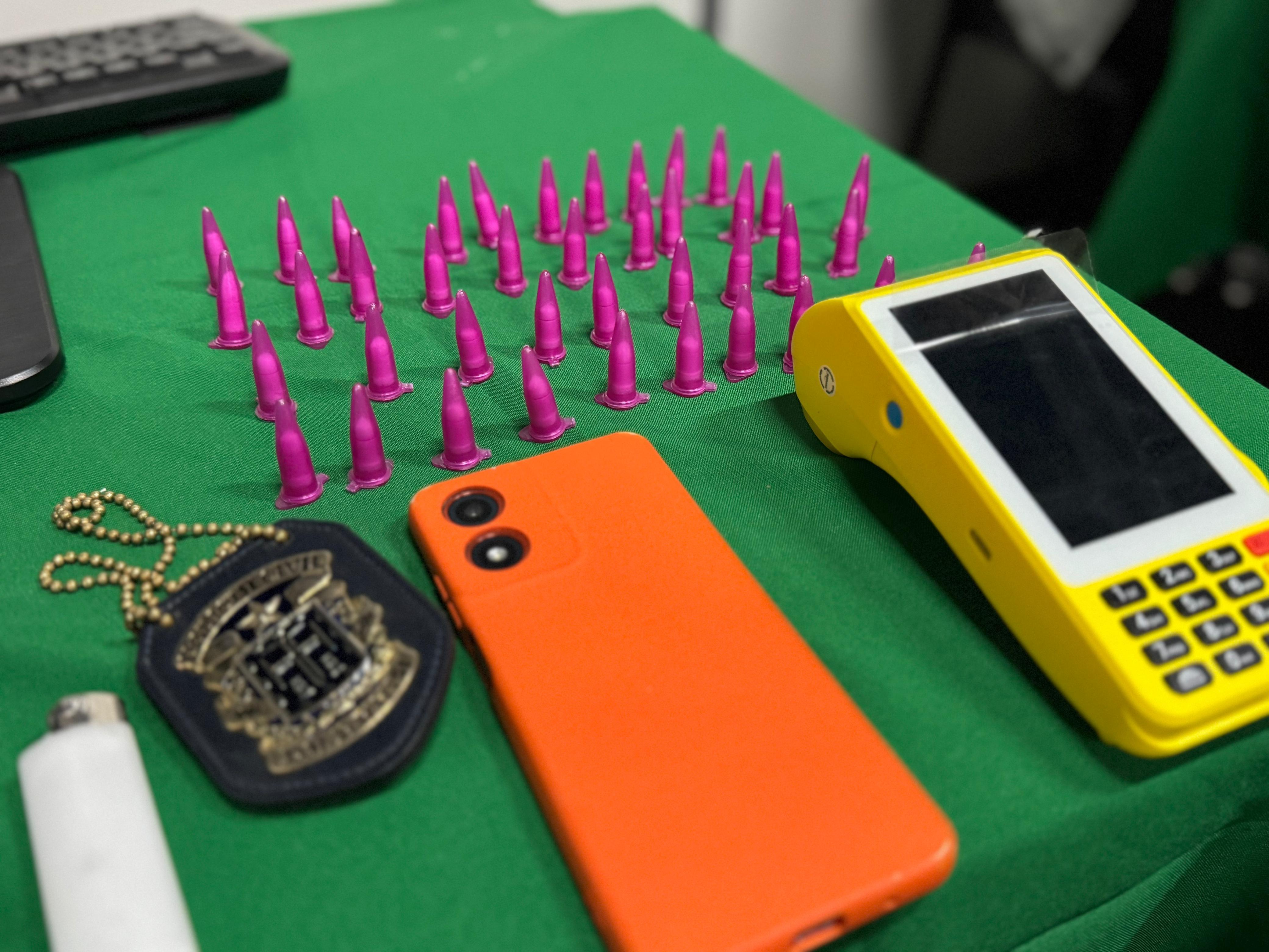 Objetos dispostos sobre uma mesa verde, incluindo embalagens pequenas de cor rosa, celular laranja, máquina de cartão amarela e distintivo policial.
