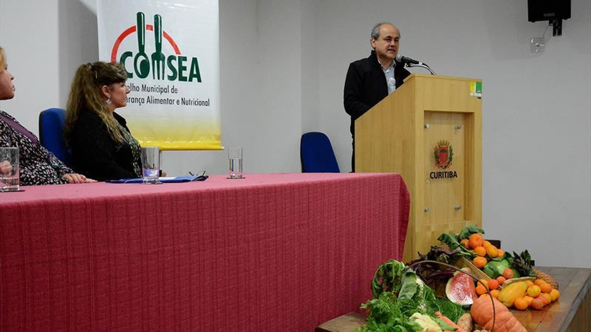 Representante discursa durante evento do Conselho Municipal de Segurança Alimentar e Nutricional de Curitiba, com participantes à mesa e alimentos expostos à frente.