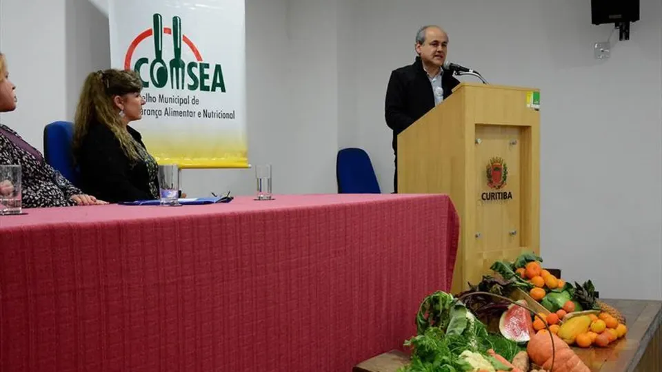 Representante discursa durante evento do Conselho Municipal de Segurança Alimentar e Nutricional de Curitiba, com participantes à mesa e alimentos expostos à frente.