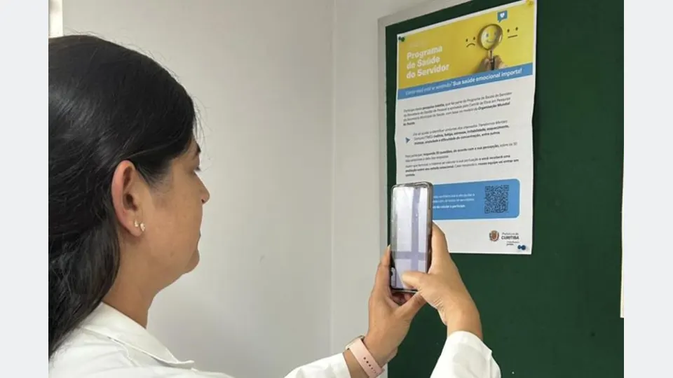Mulher utiliza celular diante de cartaz da pesquisa sobre riscos psicossociais dos servidores da Prefeitura de Curitiba.