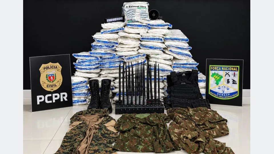 Produtos agrícolas embalados e empilhados, bloqueador de sinais com múltiplas antenas, colete tático, botas e roupas camufladas apreendidos em operação policial em Guaíra, Paraná, com símbolos da Polícia Civil e da Força Nacional ao fundo.