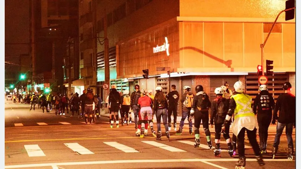 Grupo de patinadores utilizando capacetes e equipamentos de proteção participa de atividade noturna em uma rua de Curitiba.