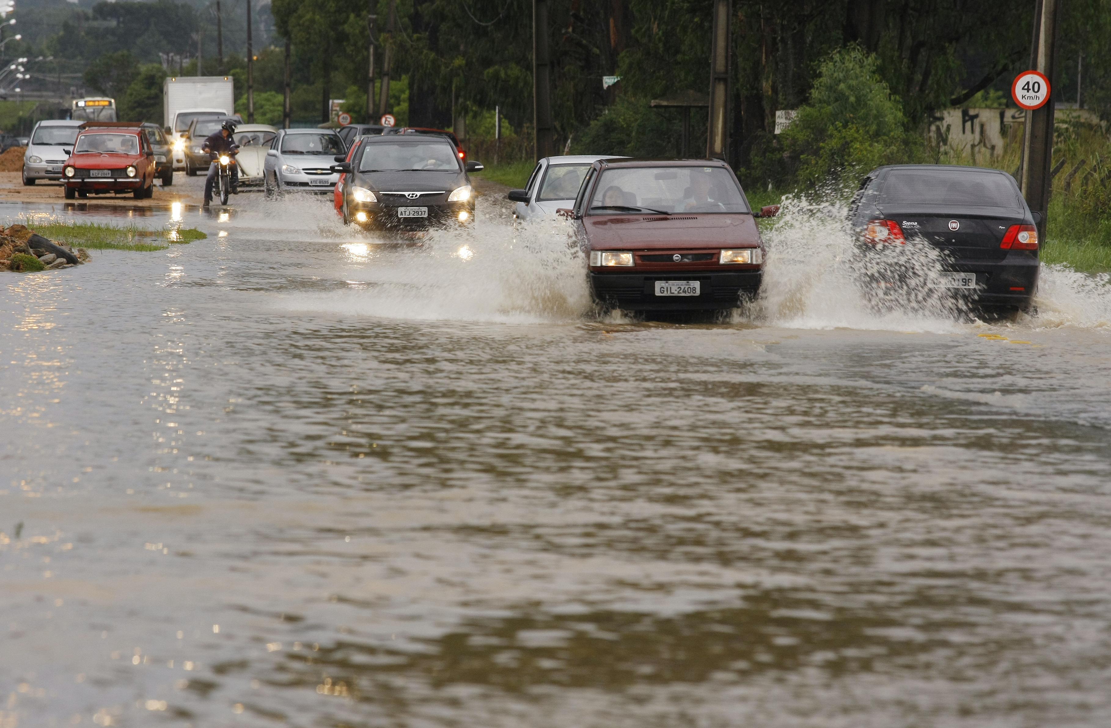 Carros e motocicleta trafegam por uma rua inundada, levantando água durante a passagem. Imagem ilustrativa de alagamentos e dificuldades no trânsito.