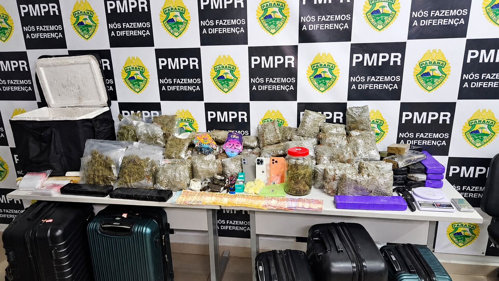 Pacotes de drogas, celulares, dinheiro, balanças e malas expostos diante de painel da Polícia Militar do Paraná após apreensão policial.