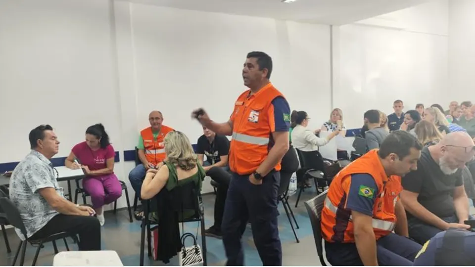 Profissional da Defesa Civil de Curitiba, usando colete laranja, participa de treinamento com servidores municipais em uma sala de capacitação.