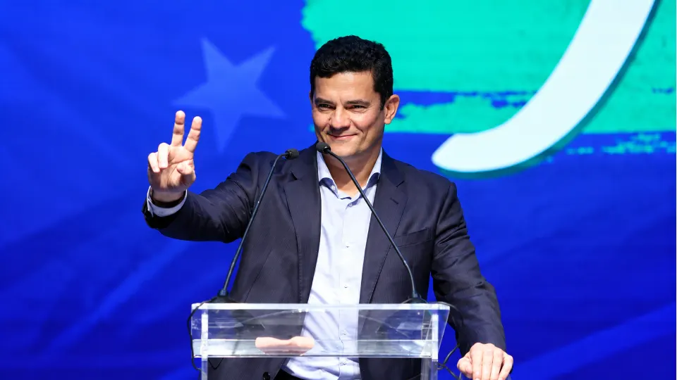 Sérgio Moro discursa em um púlpito, diante de um painel azul e verde, fazendo o sinal de vitória com a mão direita.