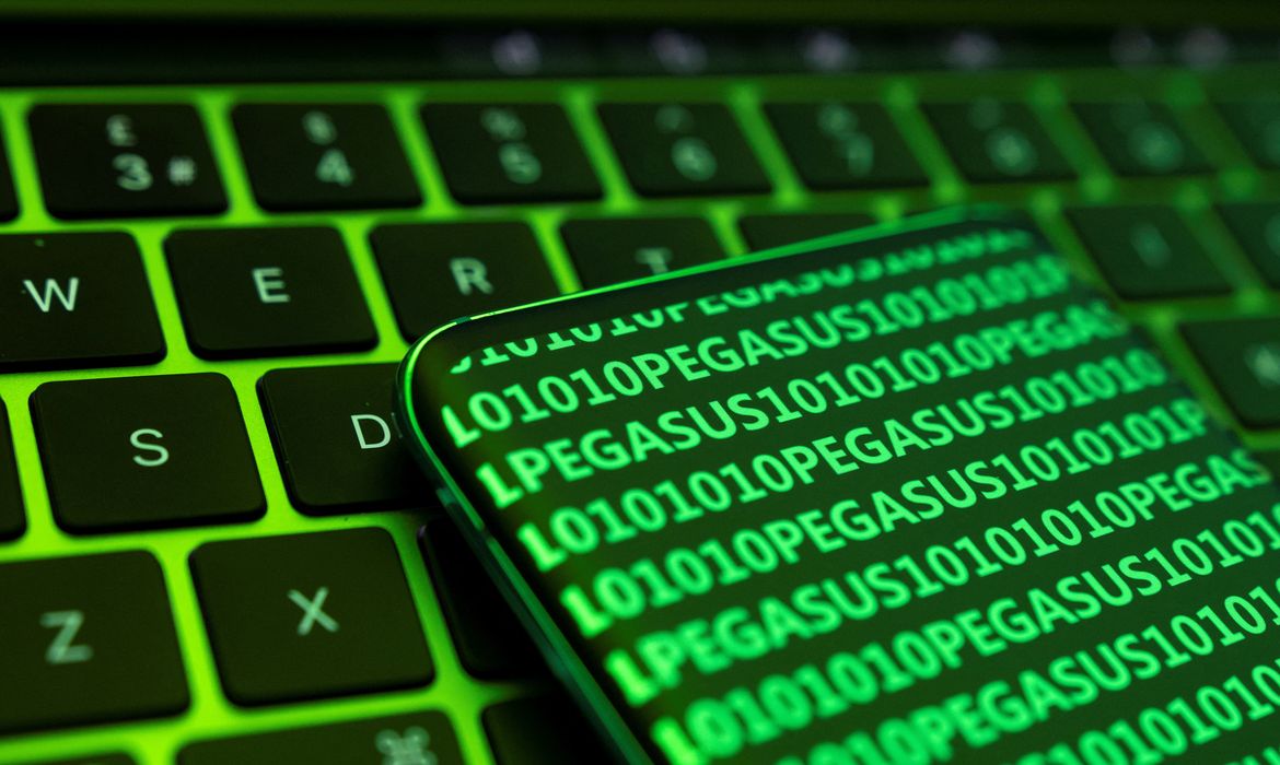 Celular sobre teclado de computador com a palavra Pegasus e códigos digitais em destaque.