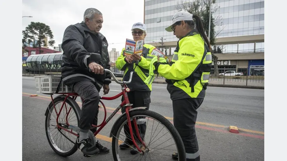 Agente de trânsito de Curitiba conversa com ciclista ao lado de uma avenida durante campanha educativa sobre segurança e circulação.