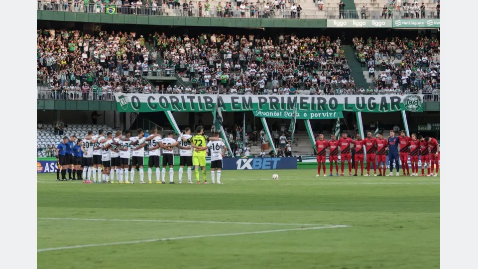 Jogadores de Coritiba e Athletico perfilados no gramado do Couto Pereira antes de clássico Atletiba, com torcida do Coritiba nas arquibancadas.
