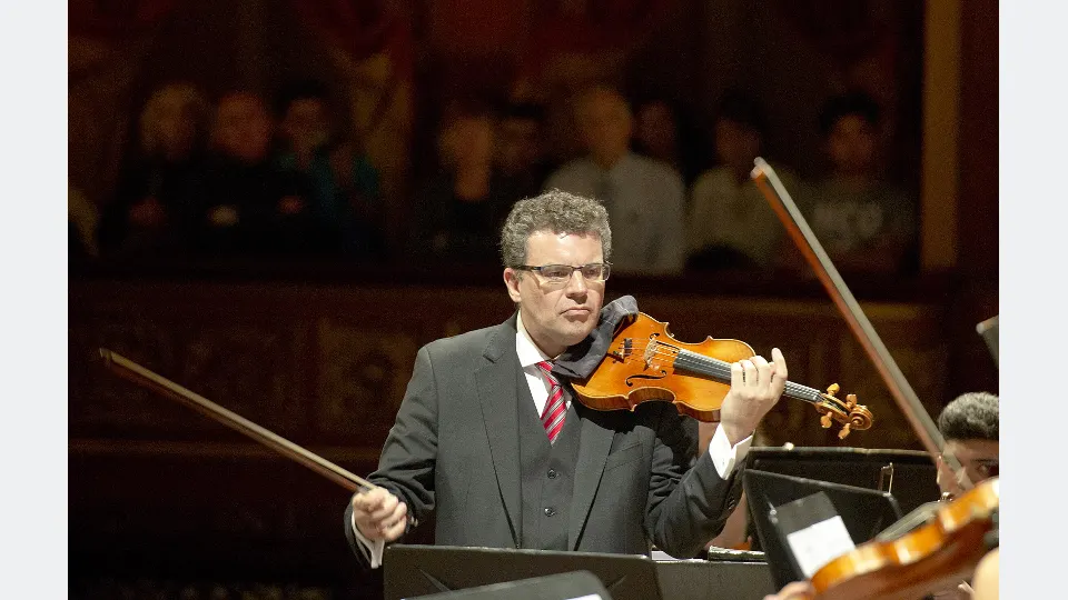 Violinista de terno executa peça musical diante da orquestra.