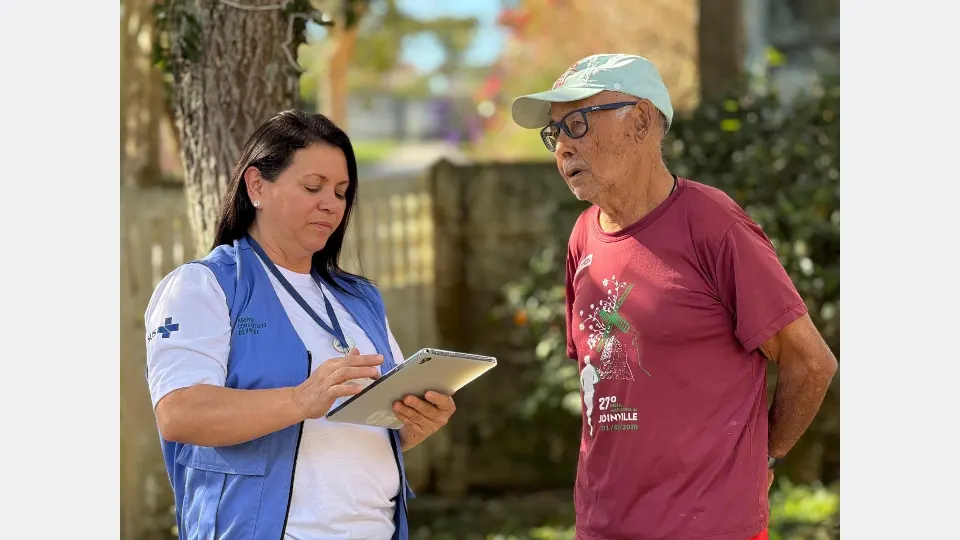 Agente comunitária de saúde com colete azul utiliza tablet durante atendimento a um homem idoso em área externa de uma residência.