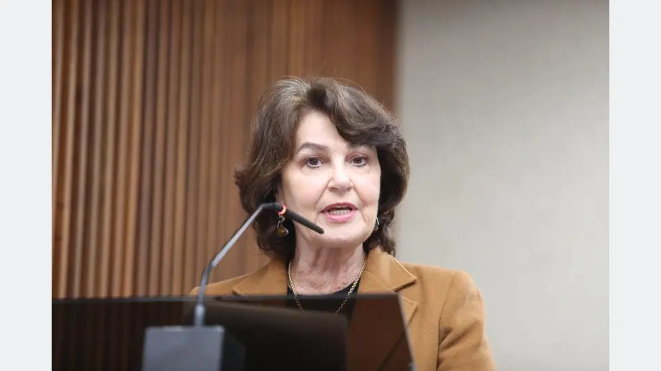 Deputada estadual Cristina Silvestri em primeiro plano, usando jaqueta marrom, com o plenário da Assembleia Legislativa do Paraná ao fundo.
