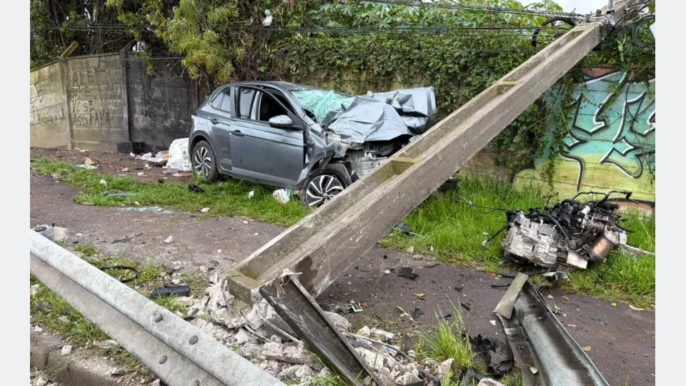 Carro cinza com a parte dianteira destruída após acidente no Boqueirão, em Curitiba. Um poste de concreto aparece caído sobre a área do veículo, enquanto o motor arrancado está no chão, próximo ao muro.