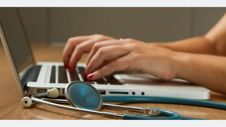 Médica digita no computador, tendo seu estetoscópio ao lado. Foto: National Cancer Institute/Unsplash